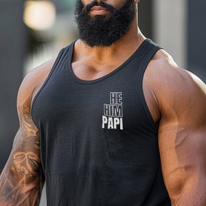 May include: A man with a full beard is wearing a black tank top with the words "HE HIM PAPI" printed on the front. He is standing in front of a blurred background.