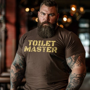 May include: A brown t-shirt with the text "TOILET MASTER" in a distressed yellow font. The shirt is worn by a person with a full beard and tattoos on their arms.