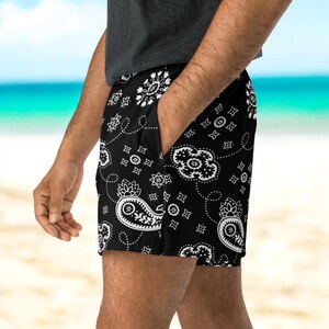 May include: Black shorts with a white paisley pattern. The shorts have a pocket and are made of a lightweight material. The shorts are worn on a beach.