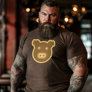 May include: A man with a brown beard and tattoos is wearing a brown t-shirt with a cartoon pig graphic. The pig is brown with yellow outlines.