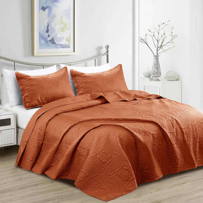 Puede incluir: Un juego de colcha de color &oacute;xido con dos fundas de almohada a juego. La ropa de cama presenta un estampado floral en relieve. La cama est&aacute; hecha con s&aacute;banas blancas. Una pintura decorativa cuelga de la pared sobre la cama.