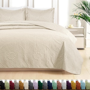 May include: A cream-colored bedspread and two pillow shams with an embossed circular pattern. The bedding is displayed on a bed with a neutral-toned wooden floor. A variety of colored bedding sets are displayed at the bottom of the image.