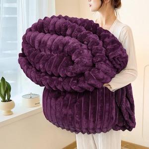 May include: A plush, deep purple blanket with a ribbed texture. The blanket is held by a person, showcasing its size and softness. The color is a rich, saturated hue, suggesting warmth and comfort. This blanket is ideal for home decor and relaxation.