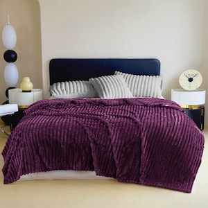 May include: A plush, deep purple blanket with a ribbed texture is draped over a bed. The blanket covers the bed and hangs over the sides. The bed has a dark blue headboard and is adorned with gray, ribbed pillows.