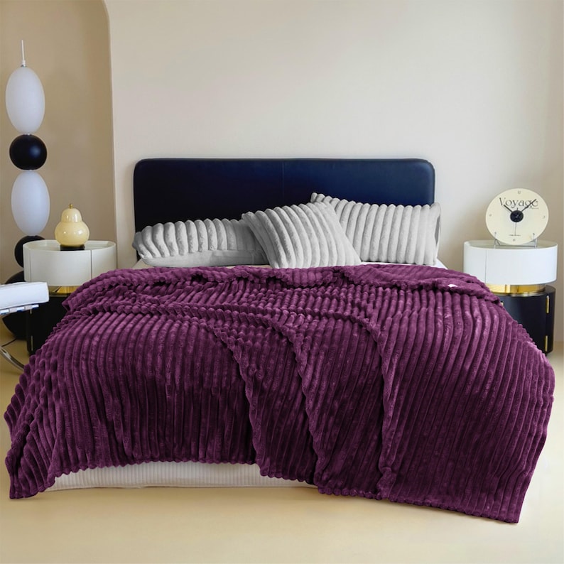 May include: A plush, deep purple blanket with a ribbed texture is draped over a bed. The blanket covers the bed and hangs over the sides. The bed has a dark blue headboard and is adorned with gray, ribbed pillows.