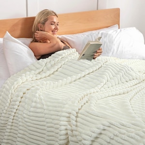 May include: A cream-colored, textured blanket with a ribbed design covers a bed. The blanket is draped over a person reading a book. The bed has white pillows and a wooden headboard. The blanket appears soft and plush.