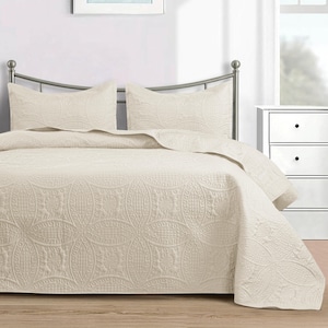 Puede incluir: Un edred&oacute;n de color crema y dos fundas de almohada a juego se muestran en una cama con estructura met&aacute;lica. La ropa de cama presenta un estampado floral en relieve. Una mesita de noche blanca es visible en el fondo.