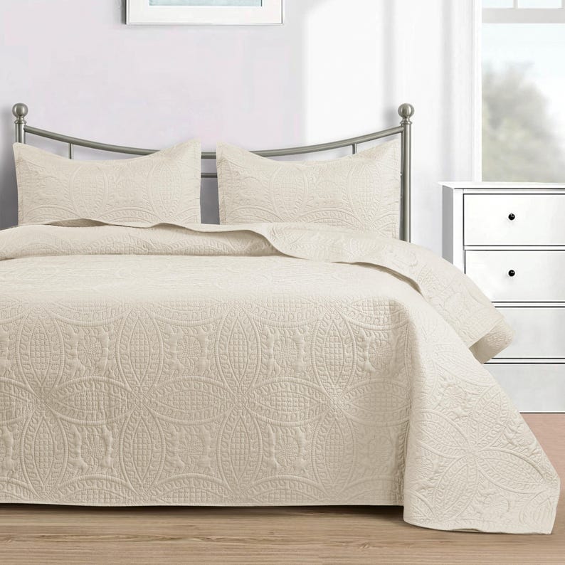 Puede incluir: Un edred&oacute;n de color crema y dos fundas de almohada a juego se muestran en una cama con estructura met&aacute;lica. La ropa de cama presenta un estampado floral en relieve. Una mesita de noche blanca es visible en el fondo.
