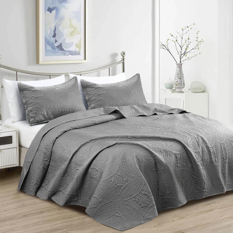 Puede incluir: Un juego de colcha gris con dos fundas de almohada a juego. La ropa de cama presenta un patr&oacute;n floral en relieve detallado. La cama est&aacute; hecha con s&aacute;banas blancas y una cabecera de metal.