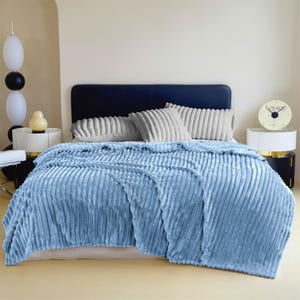 May include: A light blue, plush blanket with a ribbed texture is draped over a bed. The bed has a dark blue headboard and is adorned with matching pillows. A bedside table holds a clock and decorative items.