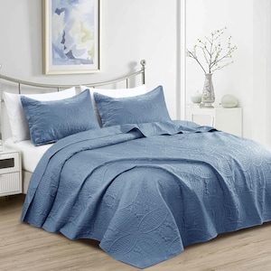 Puede incluir: Un edred&oacute;n azul y dos fundas de almohada a juego con un estampado floral en relieve. La ropa de cama est&aacute; colocada sobre una cama con cabecero plateado. La habitaci&oacute;n tiene una paleta de colores neutros con un cuadro enmarcado en la pared.