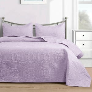 Puede incluir: Un juego de colcha de color lavanda con dos fundas de almohada a juego. La ropa de cama presenta un patr&oacute;n acolchado detallado de &oacute;valos entrelazados y dise&ntilde;os florales. El juego se muestra en una cama con cabecero de metal.