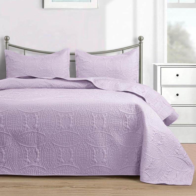 Puede incluir: Un juego de colcha de color lavanda con dos fundas de almohada a juego. La ropa de cama presenta un patr&oacute;n acolchado detallado de &oacute;valos entrelazados y dise&ntilde;os florales. El juego se muestra en una cama con cabecero de metal.
