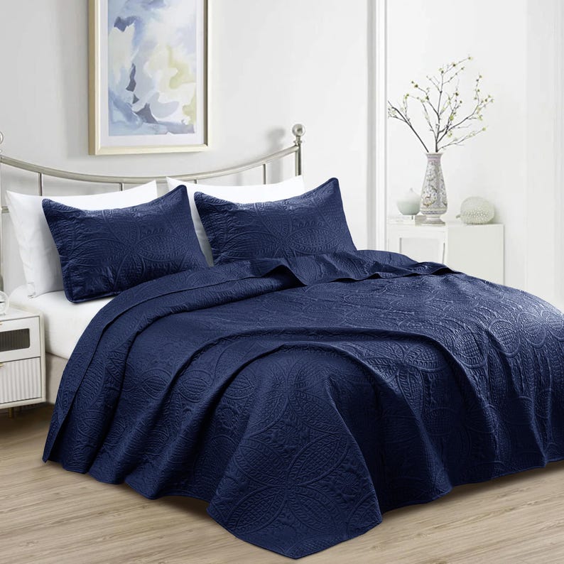 Puede incluir: Un juego de colcha azul marino con dos fundas de almohada a juego. La ropa de cama presenta un estampado floral en relieve. La cama est&aacute; hecha con s&aacute;banas blancas. Una pintura decorativa cuelga en la pared detr&aacute;s de la cama.