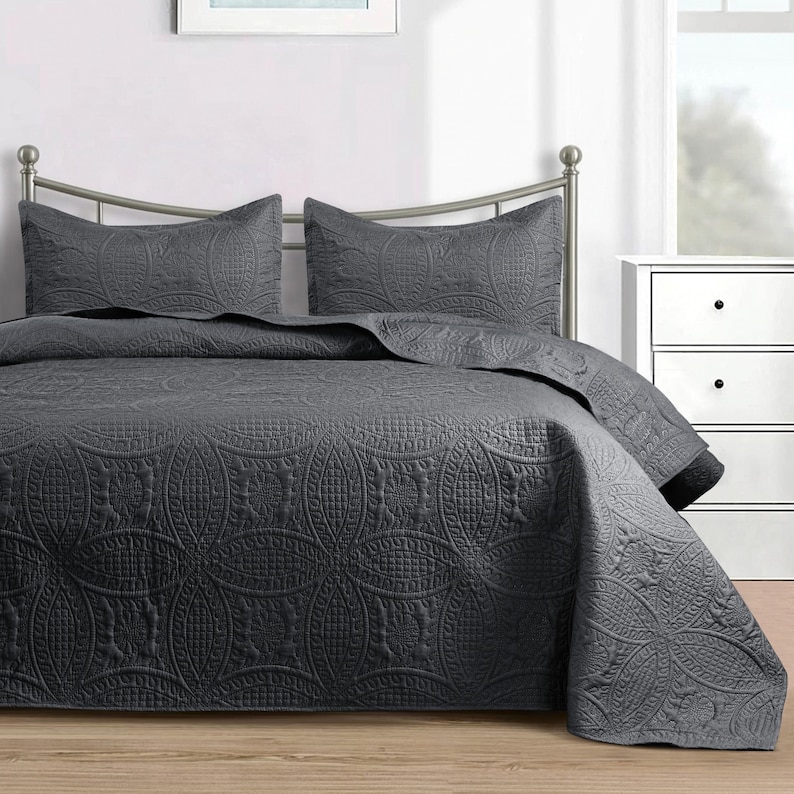 Puede incluir: Juego de colcha gris oscuro con dos fundas de almohada a juego. La ropa de cama presenta un patr&oacute;n detallado en relieve de c&iacute;rculos entrelazados y dise&ntilde;os florales. El juego se muestra en una estructura de cama de metal.