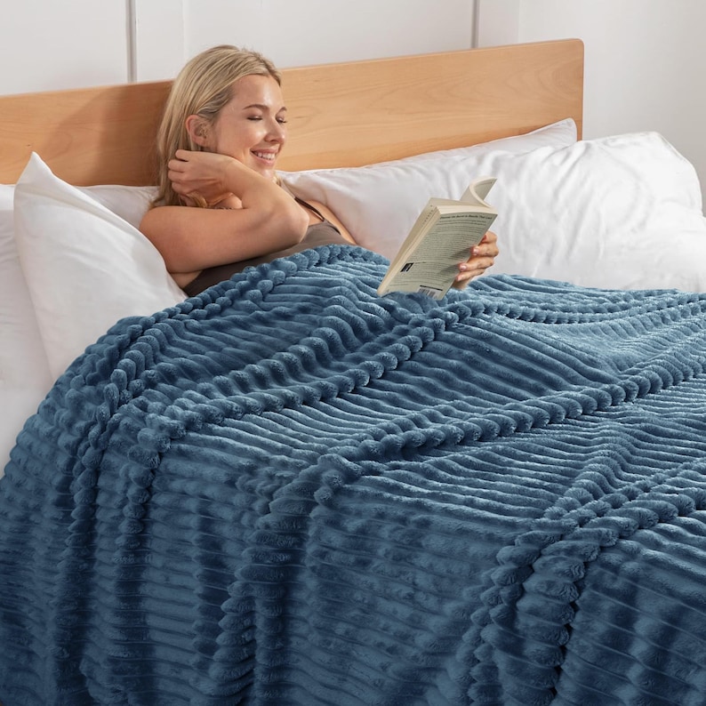 May include: A woman is relaxing in bed, covered by a textured, blue blanket. The blanket has a ribbed pattern. The woman is reading a book and smiling. The bed has white pillows and a wooden headboard.