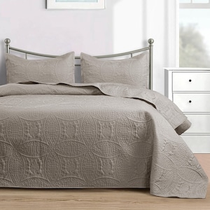 Puede incluir: Un edred&oacute;n de color topo y dos fundas de almohada con un patr&oacute;n acolchado detallado. La ropa de cama est&aacute; sobre una estructura de cama de metal en un dormitorio. El patr&oacute;n presenta c&iacute;rculos entrelazados y dise&ntilde;os florales.