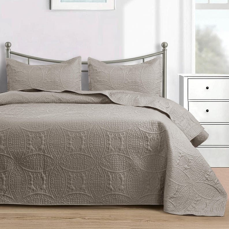 Puede incluir: Un edred&oacute;n de color topo y dos fundas de almohada con un patr&oacute;n acolchado detallado. La ropa de cama est&aacute; sobre una estructura de cama de metal en un dormitorio. El patr&oacute;n presenta c&iacute;rculos entrelazados y dise&ntilde;os florales.