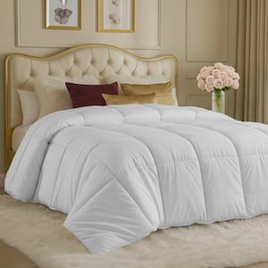 May include: A plush white comforter with a box-stitched pattern covering a bed. The bed features a tufted beige headboard, two white pillows, one dark red rectangular pillow, and two gold rectangular pillows. A vase of pink roses sits nearby.