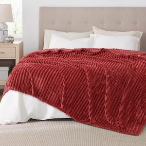 May include: A plush, brick-red blanket with a ribbed texture is draped over a bed. The blanket's diagonal pattern adds visual interest. White bedding and a neutral headboard provide a contrasting backdrop. The blanket appears soft and inviting.