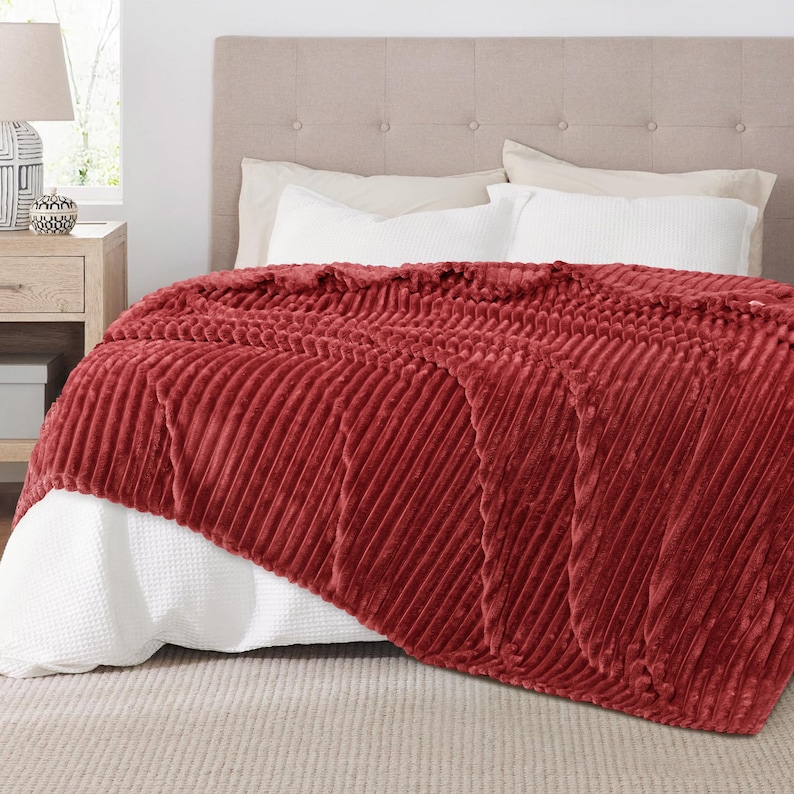 May include: A plush, brick-red blanket with a ribbed texture is draped over a bed. The blanket's diagonal pattern adds visual interest. White bedding and a neutral headboard provide a contrasting backdrop. The blanket appears soft and inviting.