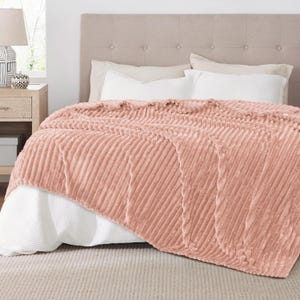 May include: A soft, blush-colored blanket with a ribbed texture is draped over a bed. The blanket's plush material contrasts with the white bedding underneath. The bed is set against a neutral-toned headboard, creating a cozy bedroom scene.