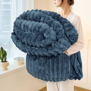 May include: A large, plush, blue blanket with a ribbed texture. The blanket is held by a person, showcasing its size and softness. The color is a deep, muted blue, suggesting warmth and comfort. The blanket appears to be made of a soft, faux fur material.