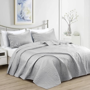 Puede incluir: Un juego de colcha y almohadas de color gris claro con un estampado floral. La ropa de cama est&aacute; cuidadosamente colocada sobre una cama con cabecero de metal. La habitaci&oacute;n est&aacute; decorada en tonos neutros, creando un ambiente relajante.