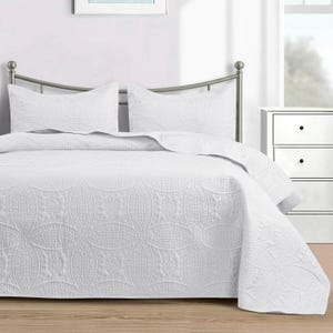 Puede incluir: Una colcha blanca y dos fundas de almohada a juego con un estampado floral en relieve. La ropa de cama est&aacute; sobre una estructura de cama de metal en un dormitorio. La colcha cubre los lados de la cama.