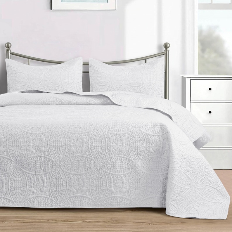 Puede incluir: Una colcha blanca y dos fundas de almohada a juego con un estampado floral en relieve. La ropa de cama est&aacute; sobre una estructura de cama de metal en un dormitorio. La colcha cubre los lados de la cama.