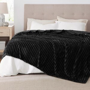 May include: A black, plush blanket with a ribbed design is draped over a bed. The blanket's texture creates a pattern of diagonal lines. The bed has white pillows and a neutral-colored headboard, creating a cozy bedroom setting.