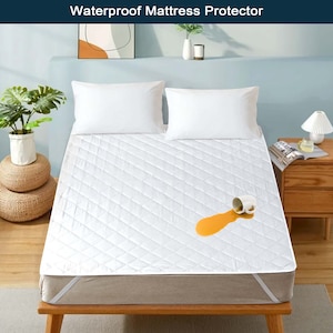 May include: A white quilted mattress protector with a diamond pattern. A white cup with a yellow liquid has spilled on the protector, showing its waterproof capabilities. The protector is on a bed frame with white sheets and pillows.