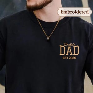 Peut inclure: T-shirt noir avec l'inscription brodée en or "Amelia's DAD EST 2026". Le t-shirt a des manches courtes et un col rond. Le mot "Embroidered" est au-dessus du texte. Un collier en or est visible.
