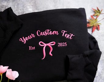 May include: Black sweatshirt with pink embroidery. The embroidery says "Your Custom Text" with a pink bow and "Est 2025".
