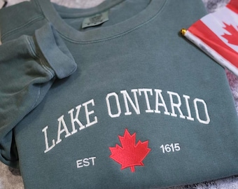 May include: A sage green crewneck sweatshirt featuring the white embroidered text LAKE ONTARIO in an arched font, with a red maple leaf graphic centered below the text, flanked by the white embroidered text EST and the number 1615.