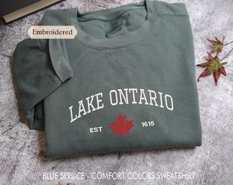 May include: A blue spruce crewneck sweatshirt featuring white embroidered text that reads LAKE ONTARIO, with the text EST 1615 and a red embroidered maple leaf centered below. A small tag labeled Embroidered is visible near the collar.