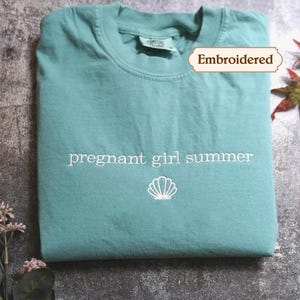 A light blue t-shirt with the text "pregnant girl summer" printed on it, along with a small white shell icon, is displayed on a gray surface. There are also some leaves and flowers in the background.