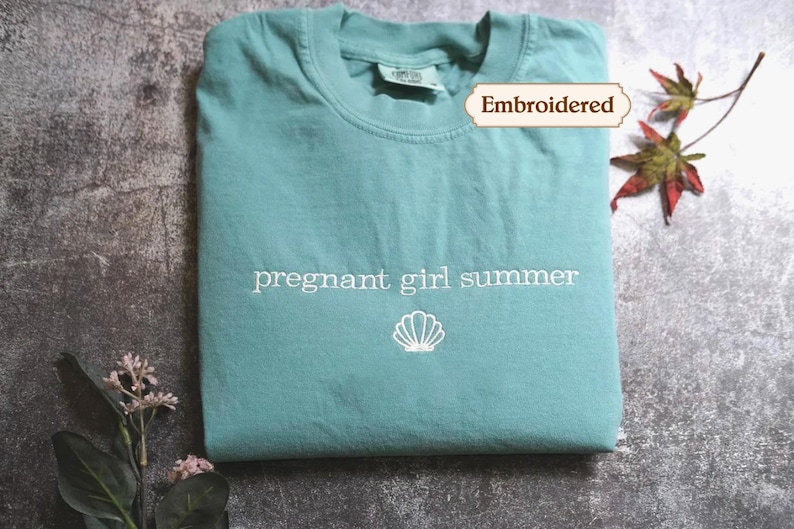 A light blue t-shirt with the text "pregnant girl summer" printed on it, along with a small white shell icon, is displayed on a gray surface. There are also some leaves and flowers in the background.