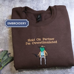 May include: A dark brown crewneck sweatshirt featuring embroidered text that reads "Hold On Partner I'm Overstimulated" above a graphic of a green frog dressed as a cowboy holding a small black pistol. The embroidery is centred on the chest.