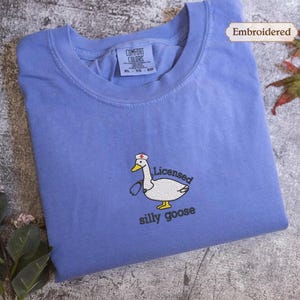 a blue t-shirt with a cartoon goose and the text "Licensed silly goose" embroidered on it. The t-shirt is placed on a gray surface, with some leaves and flowers visible in the background.