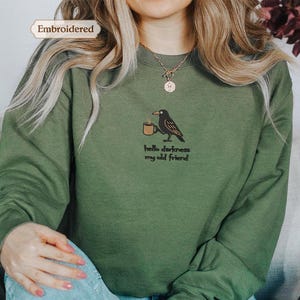 A person wearing a green sweater with a black bird embroidered on it, and the sweater has a text embroidered on it that says "Hello darkness, my old friend".