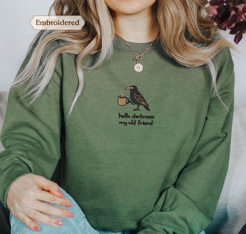 A person wearing a green sweater with a black bird embroidered on it, and the sweater has a text embroidered on it that says "Hello darkness, my old friend".