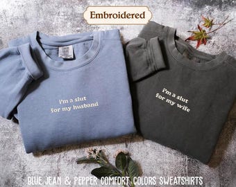 May include: Two crewneck sweatshirts in blue jean and pepper colors. The blue sweatshirt features the embroidered text 'i'm a slut for my husband' and the pepper sweatshirt features the embroidered text 'i'm a slut for my wife' in white lettering.