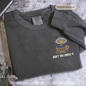 Könnte beinhalten: Pfefferfarbenes Comfort Colors Sweatshirt mit gesticktem Design. Das dunkelgraue Sweatshirt zeigt eine Kaffeetasse mit Wolken- und Blitzdesign und den Text "DON'T YOU SWEAT IT".