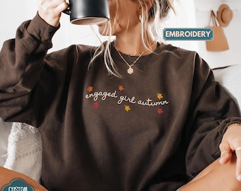 May include: A dark chocolate brown crewneck sweatshirt featuring white embroidered cursive text that reads engaged girl autumn, accented with small embroidered maple leaves in shades of orange, yellow, and red scattered around the lettering.