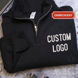 May include: Black quarter-zip sweatshirt with white embroidered "CUSTOM LOGO" text. The sweatshirt has a silver zipper and a red "EMBROIDERY" label. The garment is folded on a textured surface, with a book and leaves in the background.