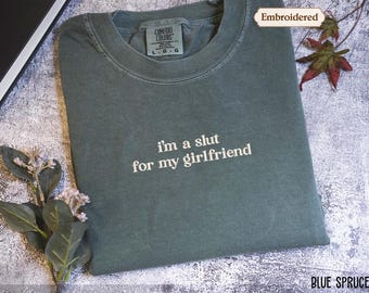 May include: A blue spruce colored crewneck t-shirt featuring white embroidered text on the chest that reads, "i'm a slut for my girlfriend." The garment has a ribbed collar and a visible interior neck label indicating a size large.