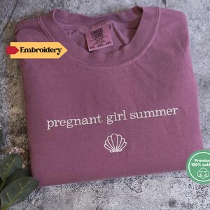 A purple t-shirt with the text "pregnant girl summer" printed on it, along with a small white shell icon, is displayed on a gray surface.