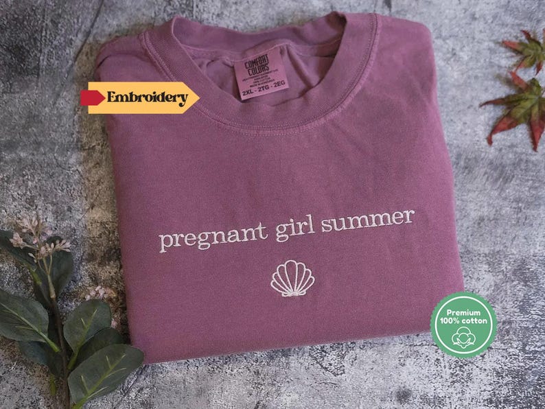 A purple t-shirt with the text "pregnant girl summer" printed on it, along with a small white shell icon, is displayed on a gray surface.