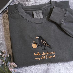 a gray sweatshirt with a black bird embroidered on it, holding a cup, and the text "Hello darkness, my old friend" printed on it.
